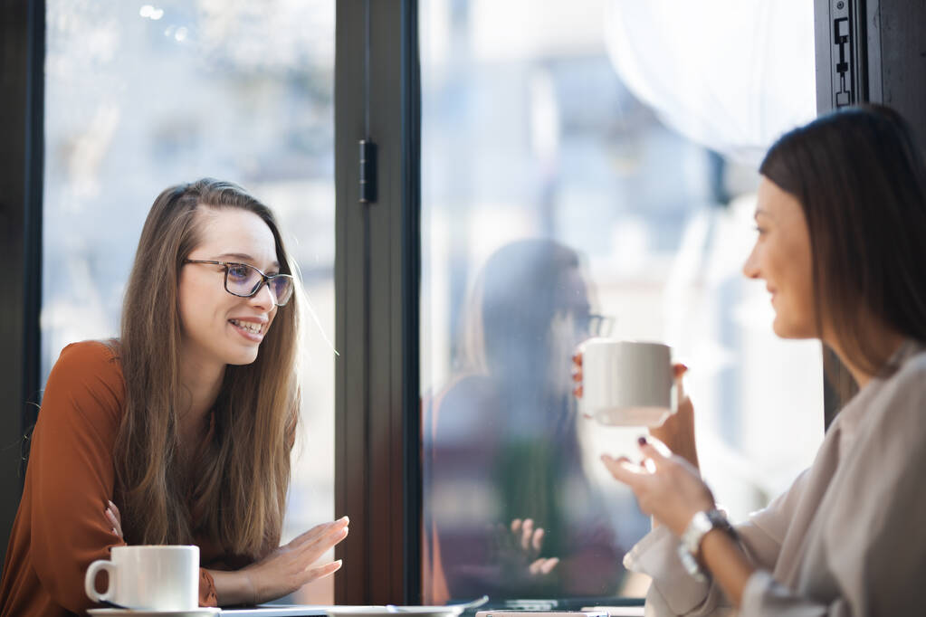 Twee jonge zakenvrouwen in een café die één op één vergadering hebben. Vrienden na het werk praten roddelen en koffie drinken aan een raam tafel op een zonnige dag. - Foto, afbeelding