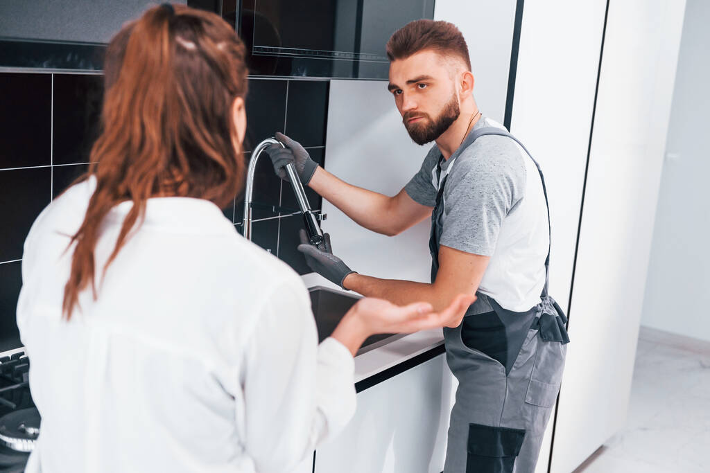 Young professional plumber in grey uniform shows results of work for housewife on the kitchen. - Foto, Bild