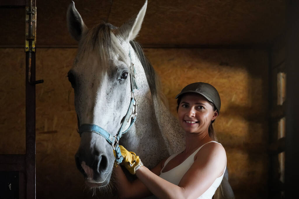 Young woman wearing riding gloves on hand standing near horse inside stables - Foto, Bild