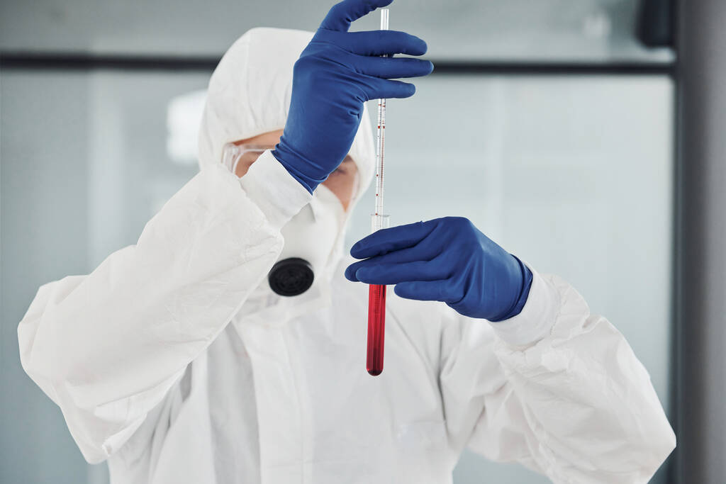 Hombre científico médico en bata de laboratorio, gafas defensivas y máscaras funciona con probeta con sangre dentro de ella. - Foto, imagen