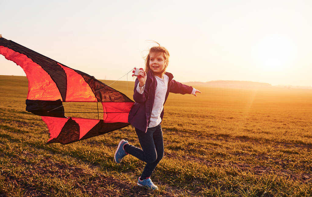 Szczęśliwa dziewczynka biegnie z latawcem w rękach na pięknym polu o świcie. - Zdjęcie, obraz