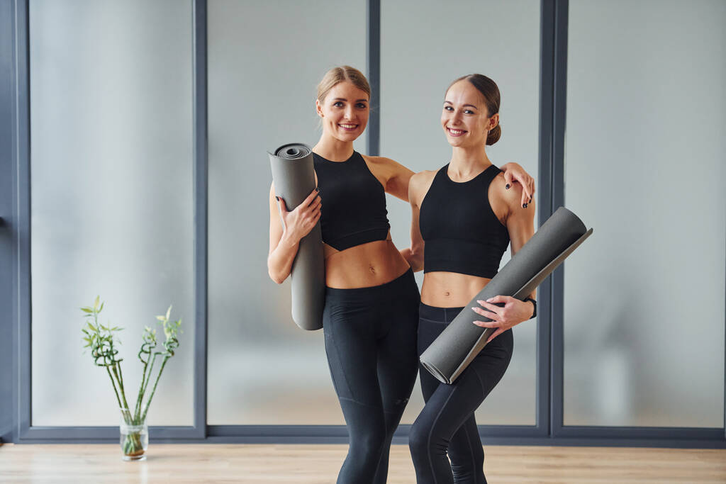 Standing with mats in hands. Two women in sportive wear and with slim bodies have fitness yoga day indoors together. - Foto, imagen