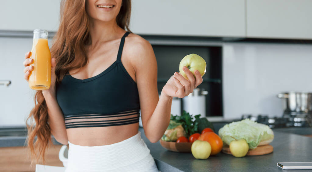 Holds apple in hand. Young european woman is indoors at kitchen indoors with healthy food. - Foto, Imagen