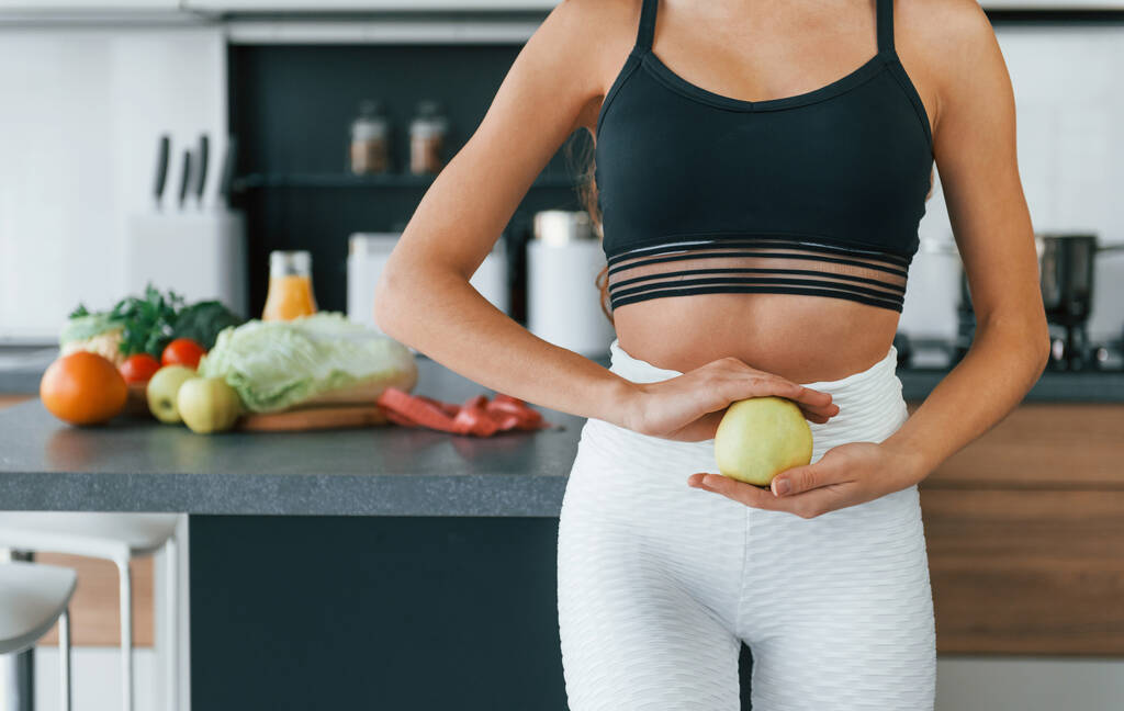 Holds apple in hand. Young european woman is indoors at kitchen indoors with healthy food. - Photo, Image