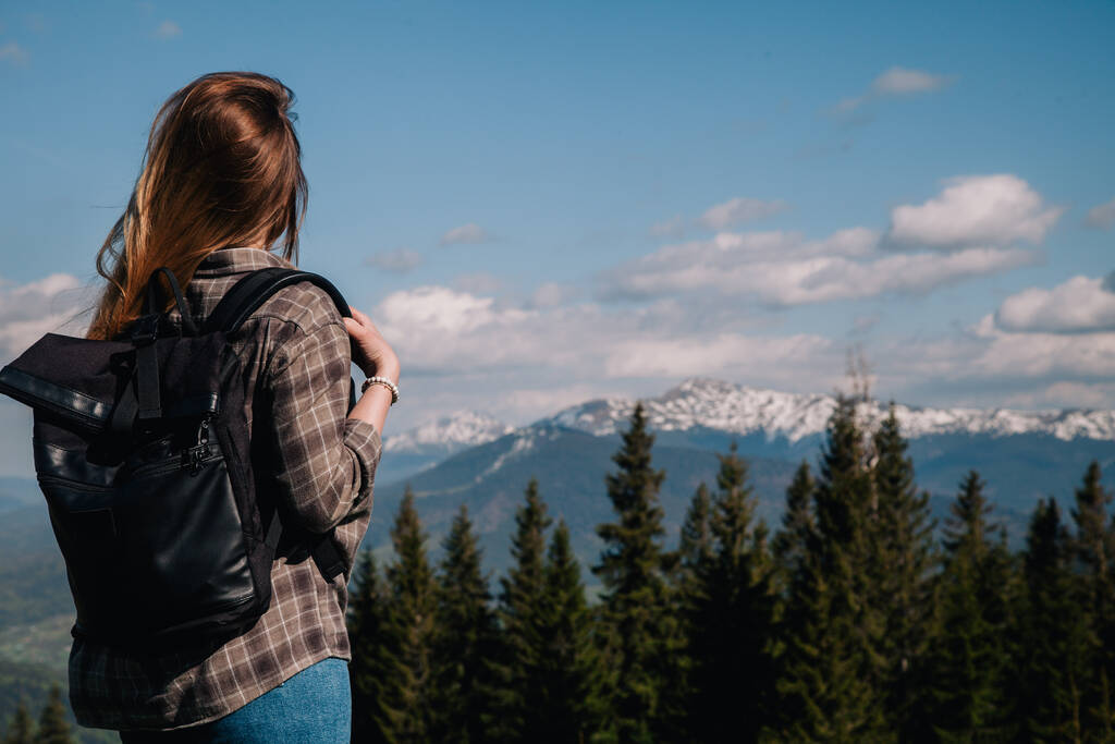 A young, slender girl with loose hair in a plaid shirt and jeans with a black backpack stands against the backdrop of mountains in sunny weather. View from the back. - Foto, afbeelding