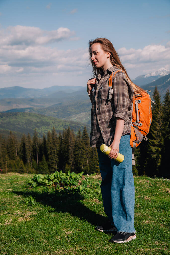 A young, girl in a plaid shirt and jeans with loose hair with an orange backpack drinks tea from a bright thermal cup against the backdrop of the mountains in the Carpathians. view from the back. - Zdjęcie, obraz
