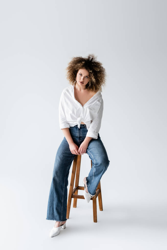 Кудрявая модель в блузке сидит на стуле на белом фоне  - Фото, изображение