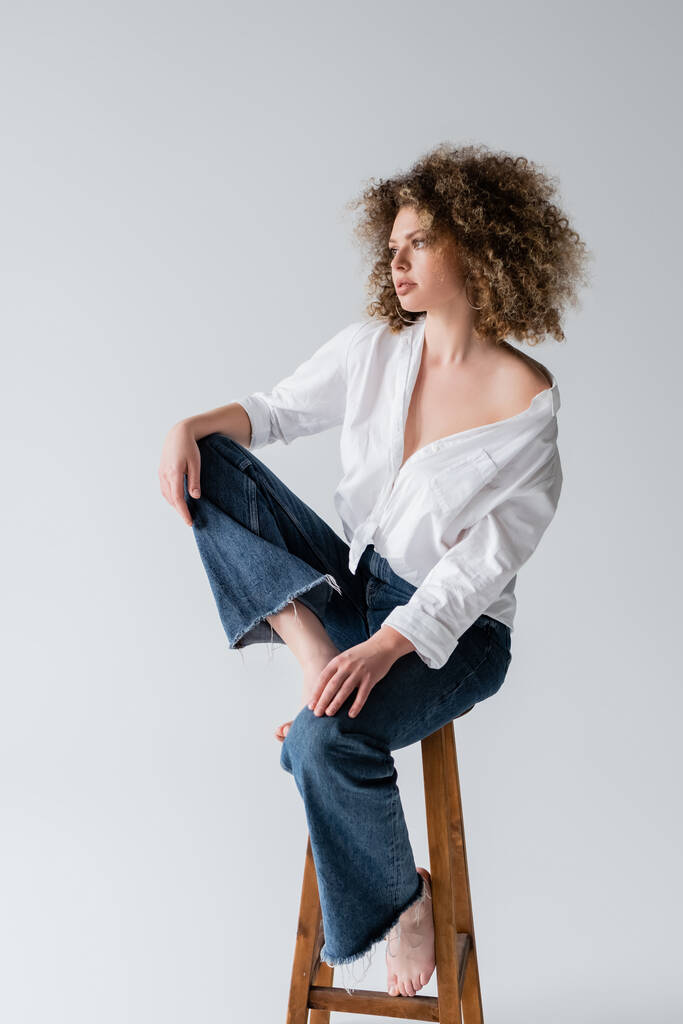 Joven modelo rizado y descalzo posando en silla aislada en blanco  - Foto, imagen