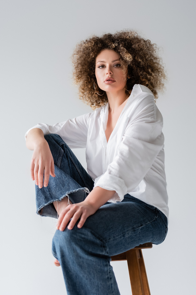 Low angle view of stylish curly woman looking at camera while sitting on chair isolated on white  - Photo, Image