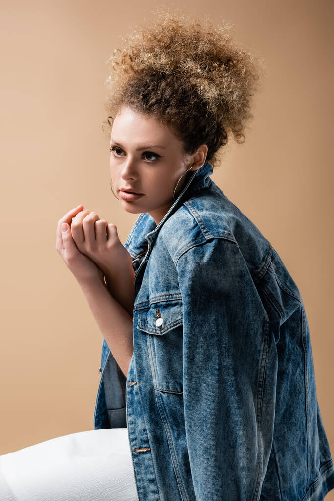 Stylish curly woman in denim jacket touching hands isolated on beige  - Photo, Image