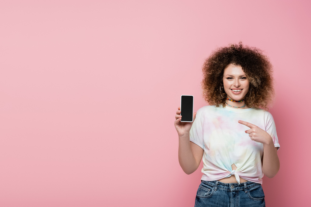 Mujer alegre señalando el teléfono móvil aislado en rosa  - Foto, imagen
