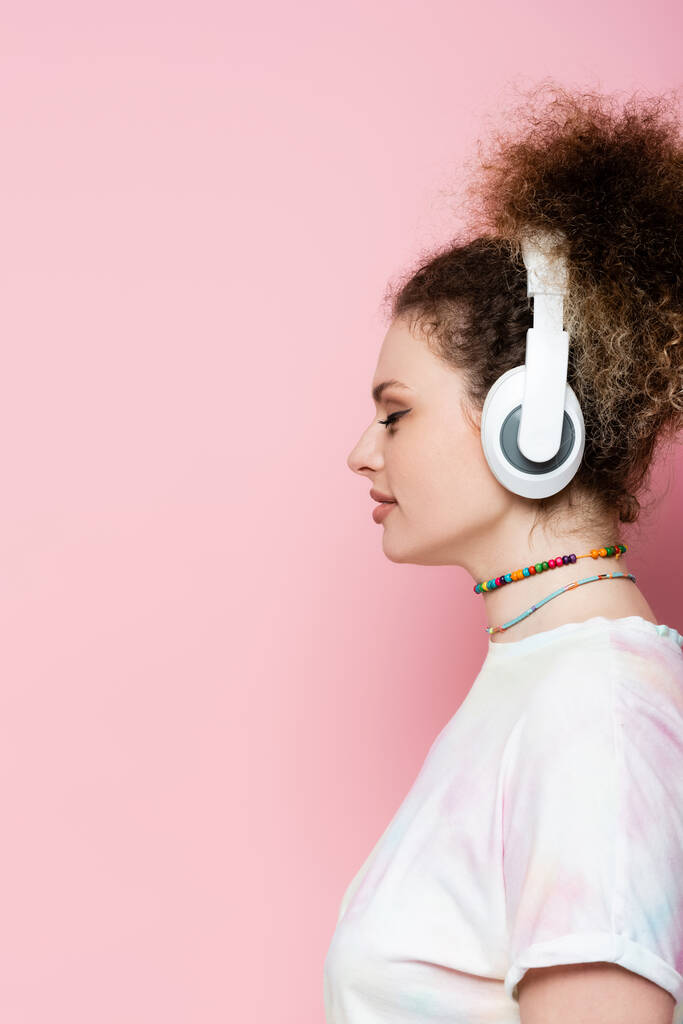 Vista lateral da mulher encaracolada em fones de ouvido sem fio no fundo rosa - Foto, Imagem