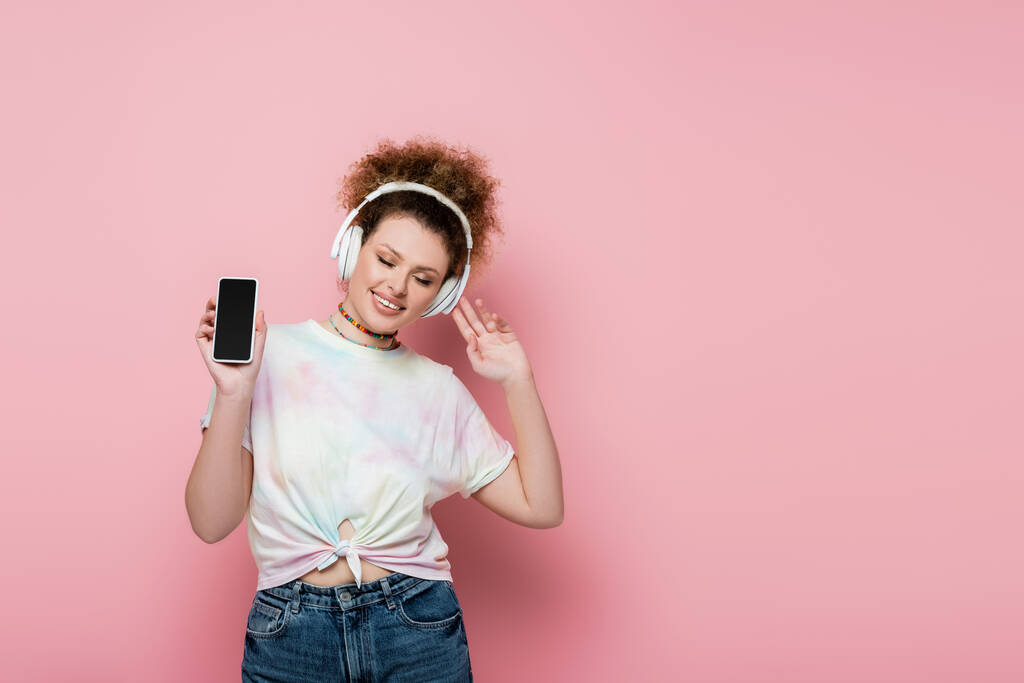 Curly woman in headphones holding cellphone on pink background - Photo, Image