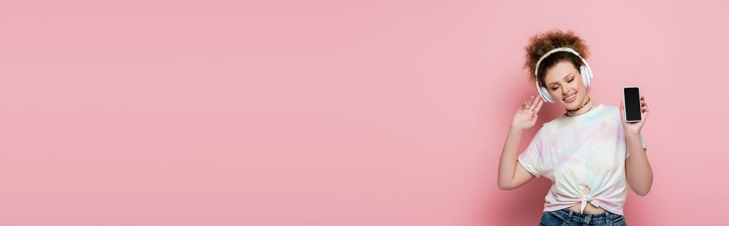 Веселая женщина в наушниках показывает смартфон на розовом фоне, баннер  - Фото, изображение