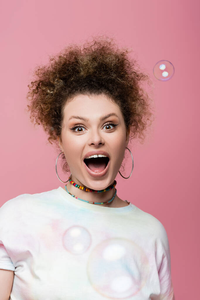 Excited curly woman looking at camera near soap bubbles on pink background - Photo, Image
