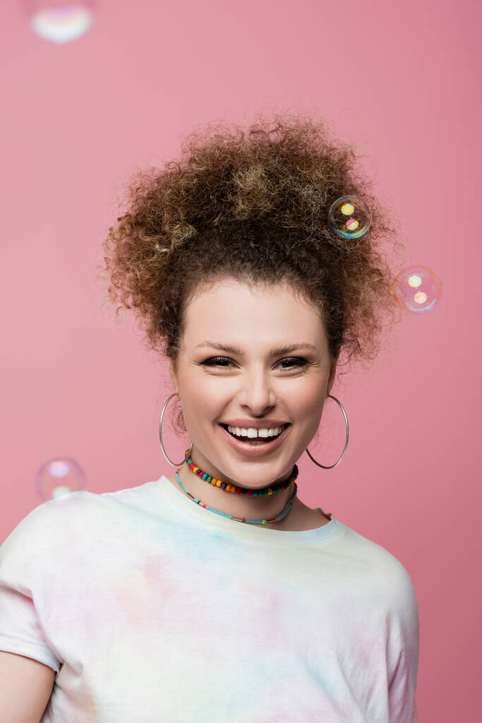 Positive woman looking at camera near soap bubbles on pink background - Photo, Image