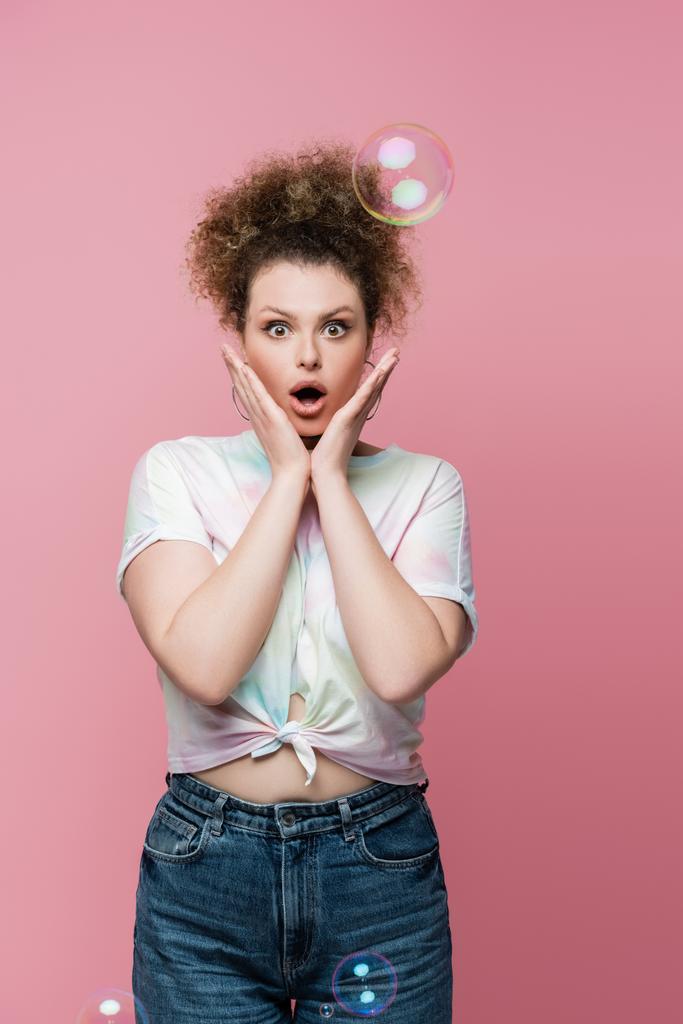 Geschokte vrouw in jeans en t-shirt op zoek naar camera in de buurt van zeepbellen op roze achtergrond - Foto, afbeelding