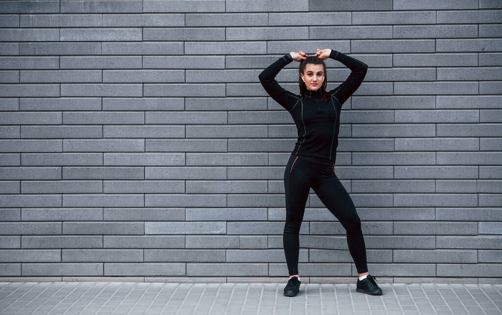 Young sportive girl in black sportswear standing outdoors near gray wall. - Photo, Image