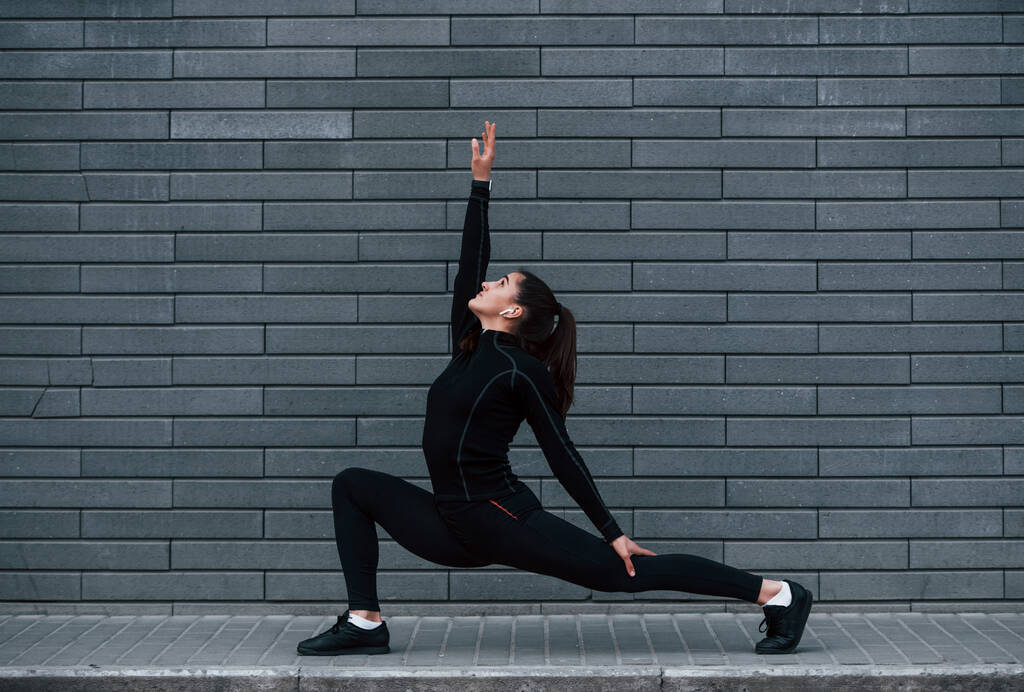 Young sportive girl in black sportswear outdoors doing yoga exercises near gray wall. - Foto, afbeelding