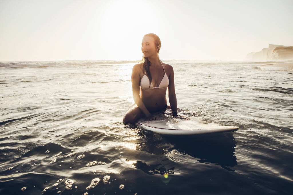 Sörf tahtasının üzerinde oturmuş dalgaları bekleyen güzel genç bir kadın. - Fotoğraf, Görsel