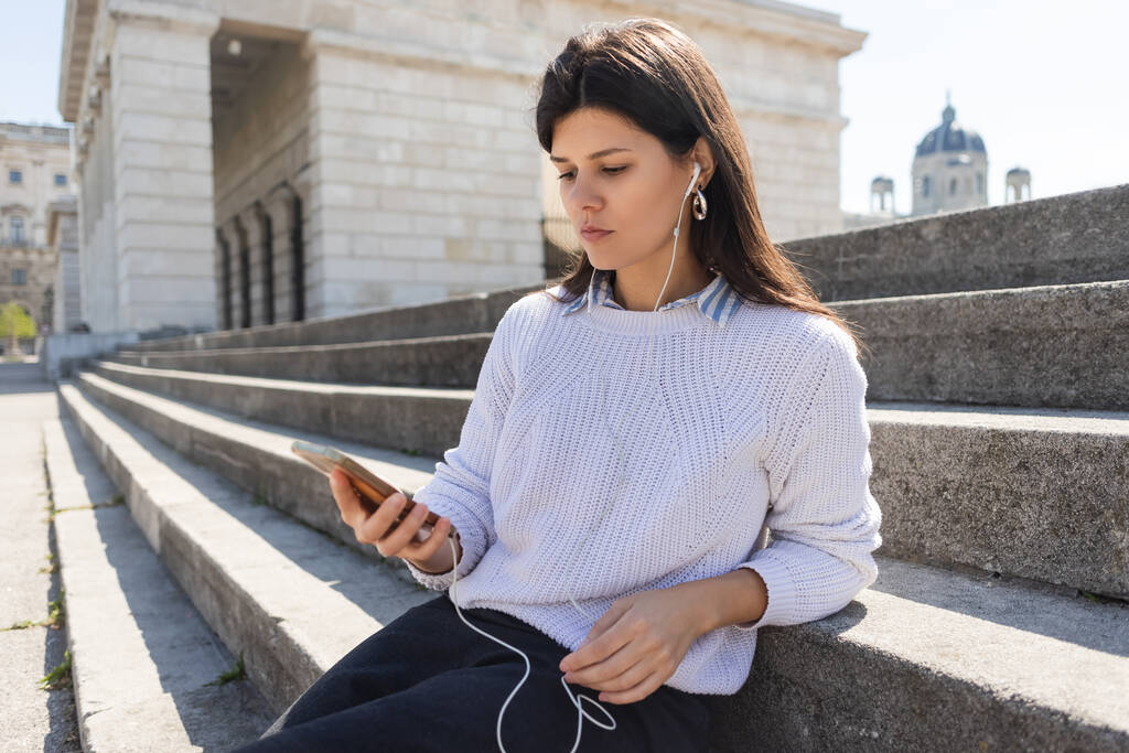 brünette Frau hört Musik in kabelgebundenen Kopfhörern und benutzt Smartphone, während sie auf der Treppe sitzt  - Foto, Bild