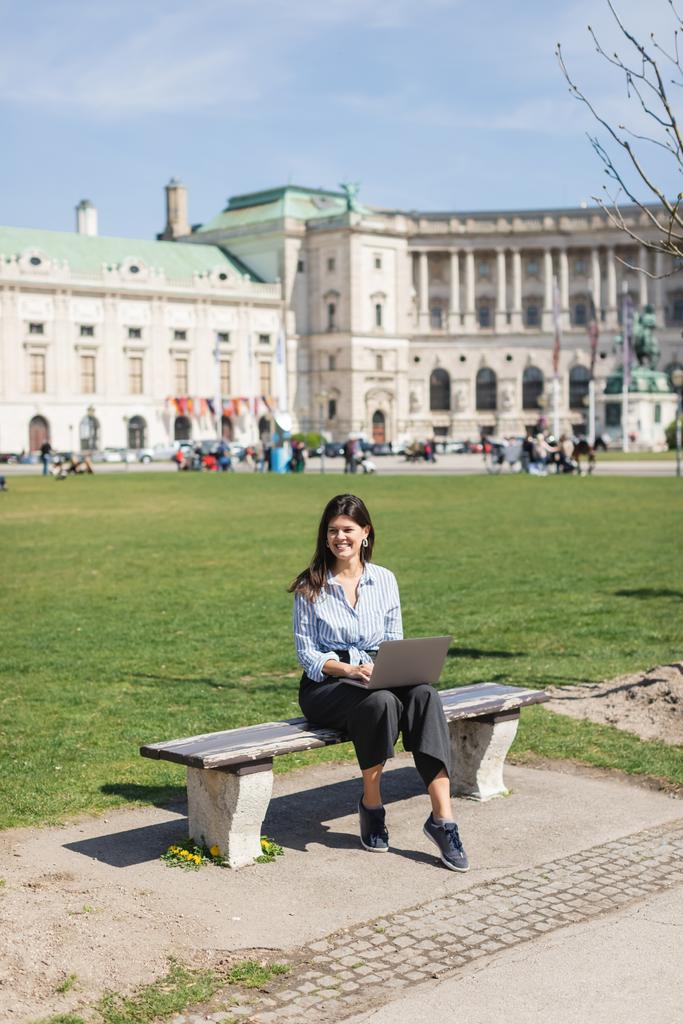 glückliche Frau auf Bank sitzend und mit Laptop im grünen Park von Wien - Foto, Bild