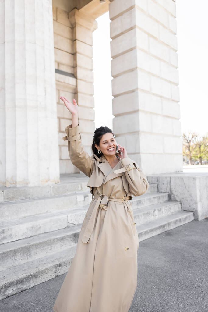 ベージュトレンチコートを着た幸せな女性が手を振っている間にスマートフォンで話している間ウィーンの建物の近くの通り - 写真・画像