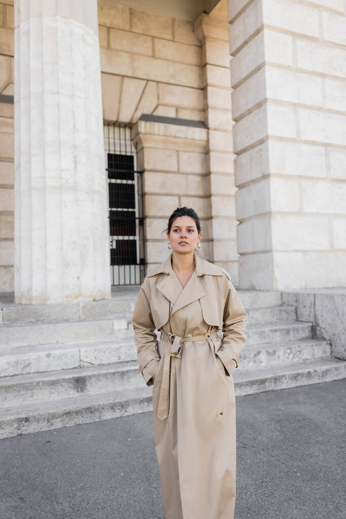 brunette woman in beige trench coat looking at camera while standing with hands in pockets near building of vienna - Photo, Image