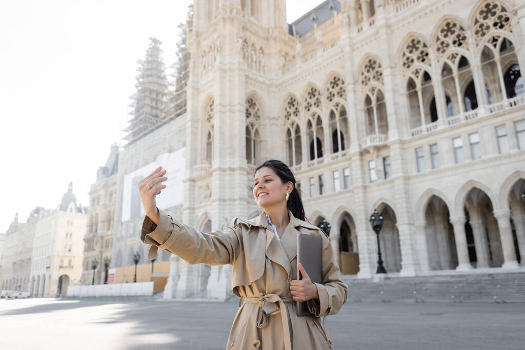freelancer in beige trench coat taking selfie while holding laptop near vienna city hall  - Photo, Image