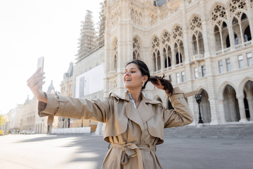 Glückliche Frau im beigen Trenchcoat macht Selfie in der Nähe des Wiener Rathauses - Foto, Bild