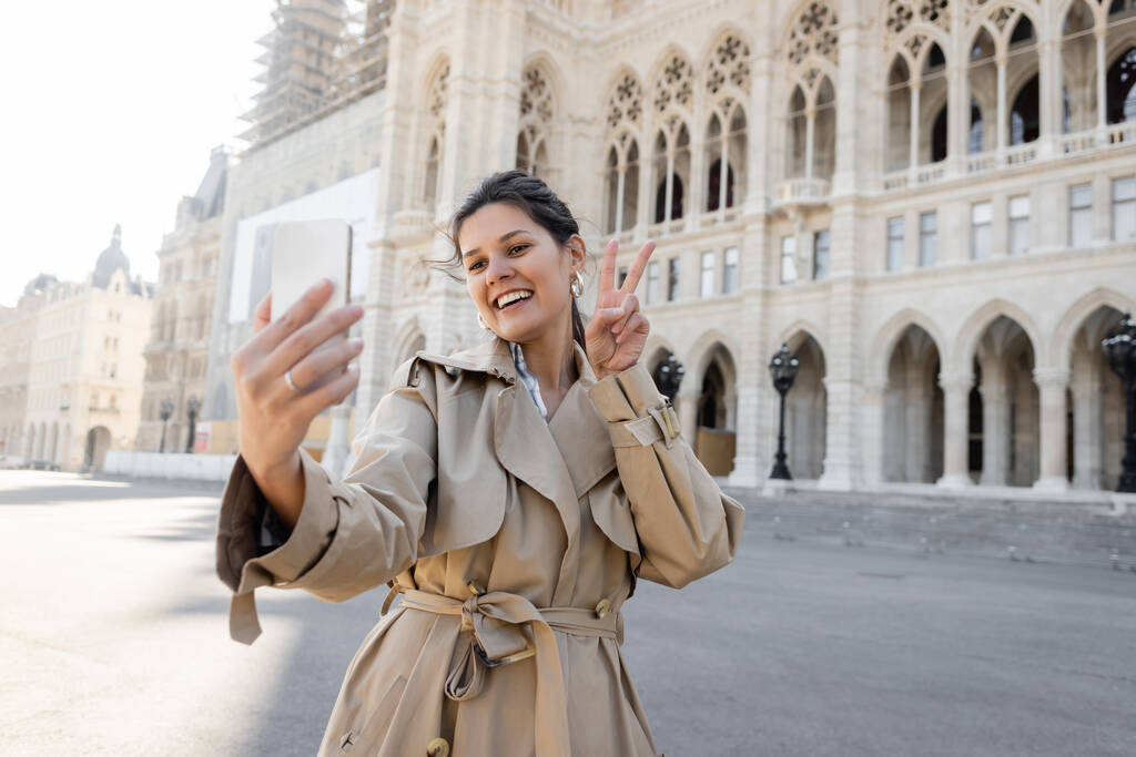 happy woman in beige trench coat showing peace sign taking selfie near vienna city hall - Photo, Image