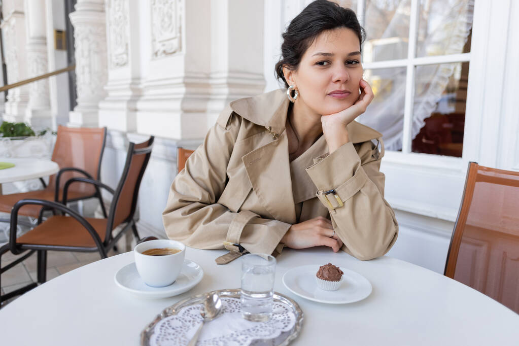 брюнетка в пальто сидит рядом с кексом и чашкой кофе на террасе кафе - Фото, изображение