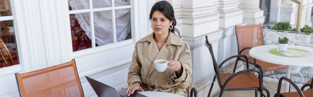 kobieta w płaszczu za pomocą laptopa i trzymając filiżankę w kawiarni taras na ulicy europejskiej w Wiedniu, sztandar - Zdjęcie, obraz