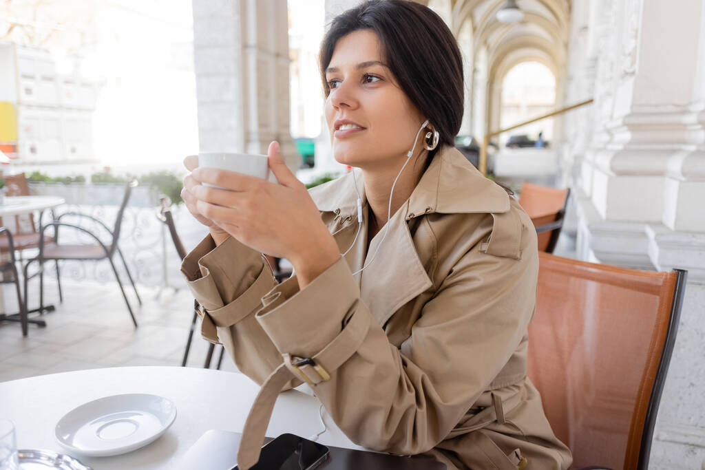 mujer feliz en gabardina escuchando música y sosteniendo la taza cerca de teléfono inteligente y portátil en la terraza de la cafetería - Foto, imagen