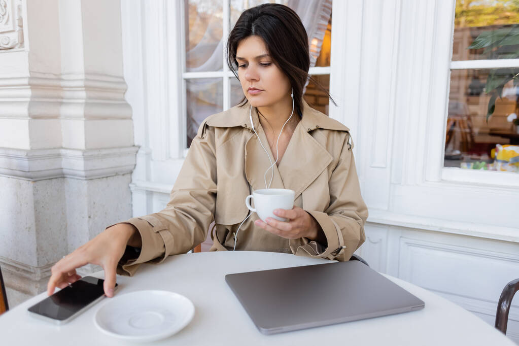 brunetta donna in trench ascoltare musica e tenere la tazza mentre raggiunge smartphone sul tavolo in terrazza caffè - Foto, immagini