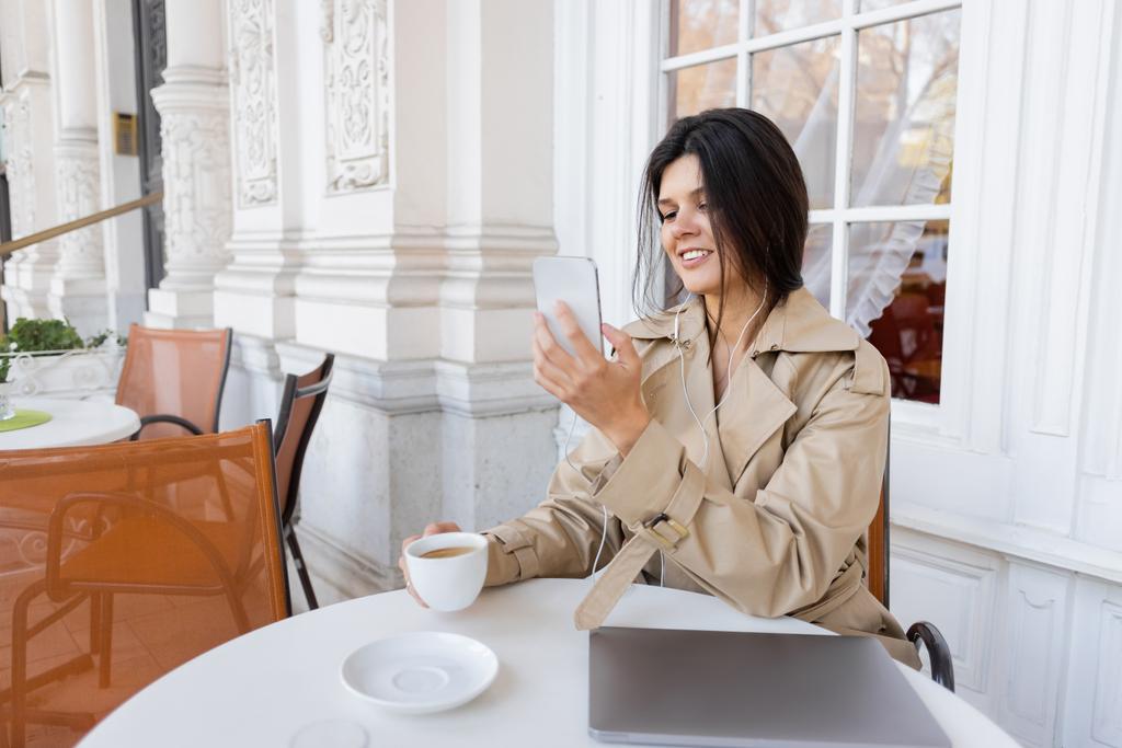 Lächelnde Frau im Trenchcoat hört Musik und hält Tasse in der Hand, während sie auf der Caféterrasse ein Foto mit dem Smartphone macht - Foto, Bild