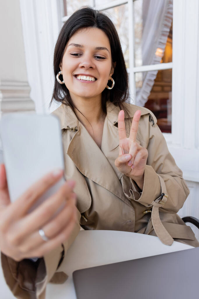 veselá žena v trenč kabát ukazující mír znamení a přičemž selfie na rozmazané smartphone v kavárně terasa - Fotografie, Obrázek