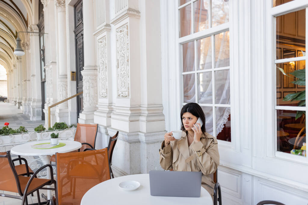 pigiste en trench coat parlant sur téléphone portable près d'un ordinateur portable et tenant une tasse de café sur la terrasse du café à Vienne - Photo, image