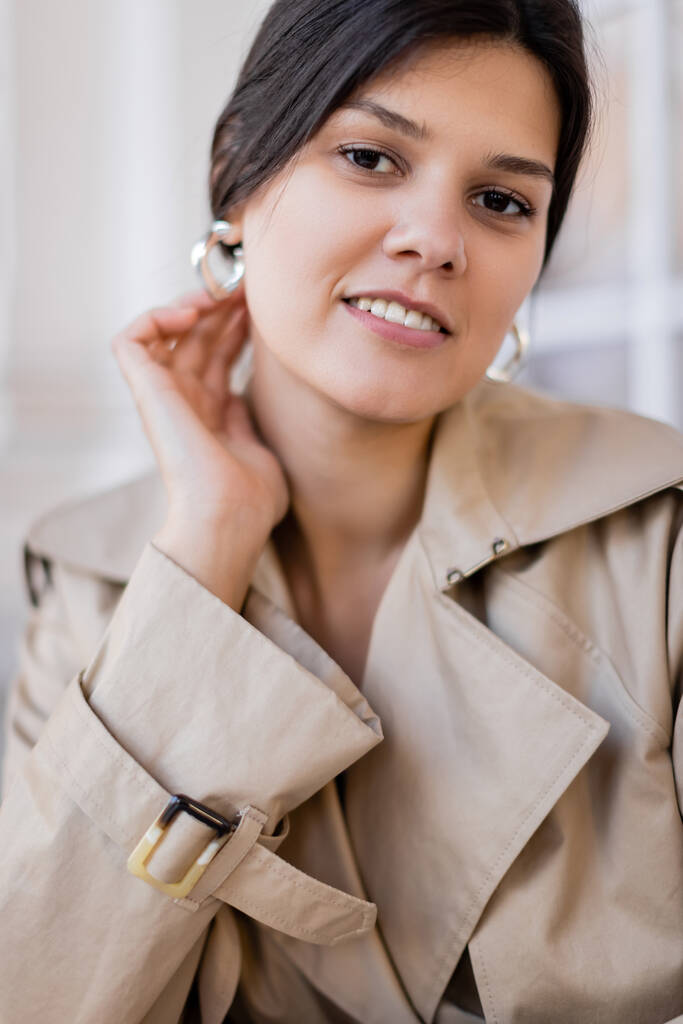 portrait de femme heureuse en trench coat beige et boucles d'oreilles cerceau souriant à l'extérieur  - Photo, image