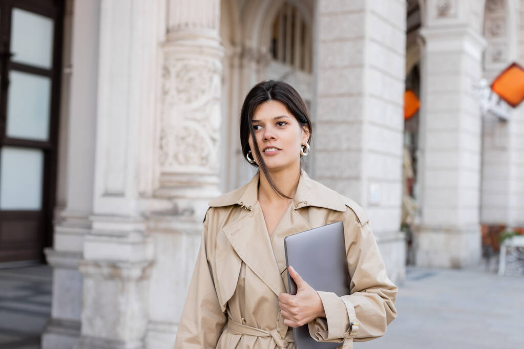 stijlvolle vrouw in beige trench jas wandelen met laptop op straat van vienna  - Foto, afbeelding