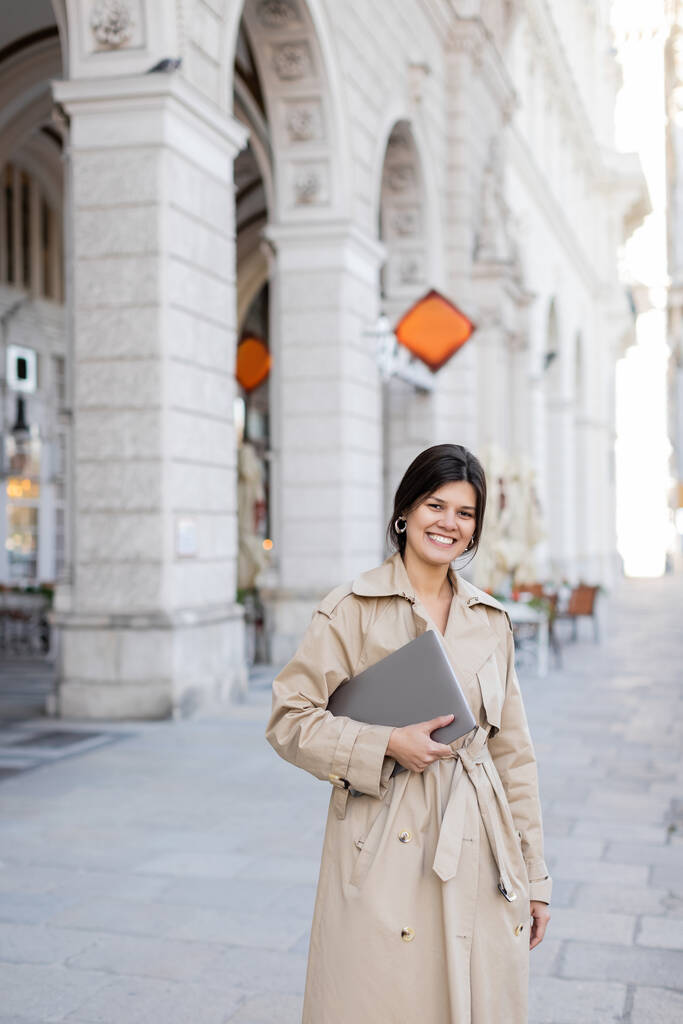 šťastná žena v trenč kabát chůze s notebookem na ulici Vídně  - Fotografie, Obrázek