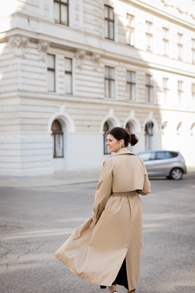 felice donna in cuffia cablata e trench che cammina per strada a Vienna - Foto, immagini