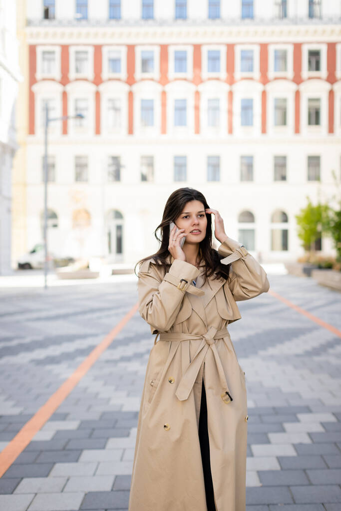 μελαχρινή γυναίκα σε μπεζ παλτό μιλώντας στο smartphone στο δρόμο της Βιέννης  - Φωτογραφία, εικόνα