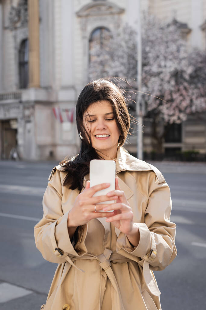Avrupa caddesinde şık ceketli neşeli bir kadın akıllı telefon kullanıyor.  - Fotoğraf, Görsel