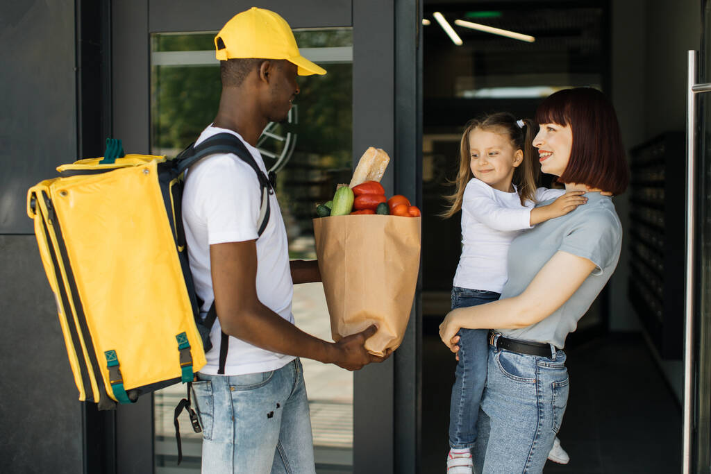 Hombre de entrega afroamericano en uniforme amarillo entrega bolsa ecológica de papel con comida para llevar de comestibles - Foto, Imagen