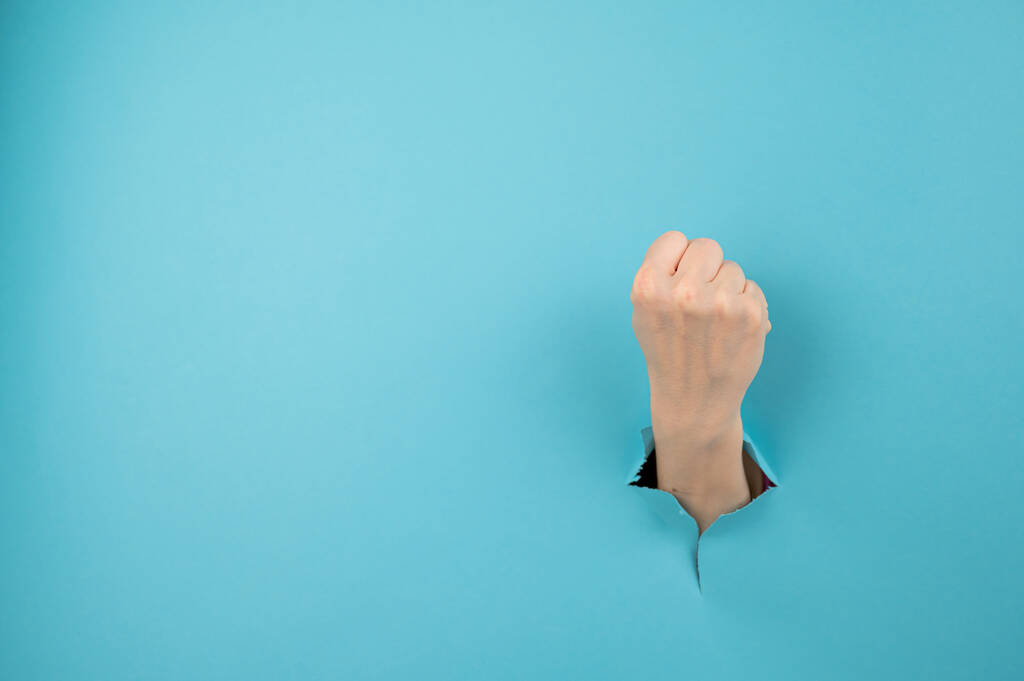 Una mano di donna che sporge da un buco su uno sfondo blu mostra un pugno. - Foto, immagini