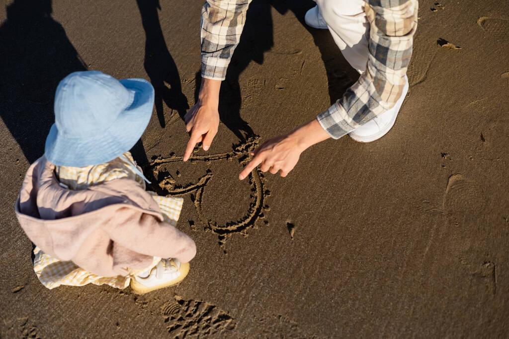 Высокий угол зрения мужчины, рисующего сердечный знак на песке рядом с маленькой дочкой  - Фото, изображение