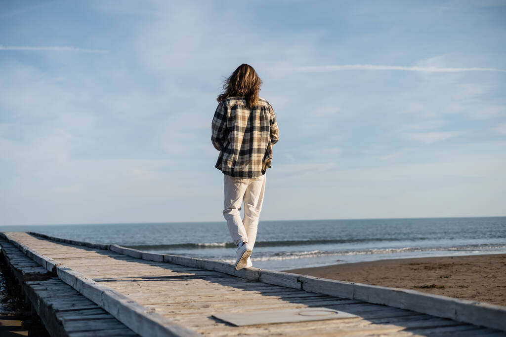 Vista posteriore dell'uomo dai capelli lunghi che cammina sul molo di legno vicino al mare Adriatico in Italia  - Foto, immagini