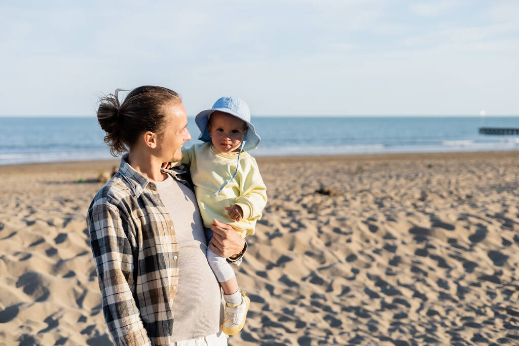 Padres positivos sosteniendo bebé en sombrero de panama en la playa cerca del mar en Italia   - Foto, imagen