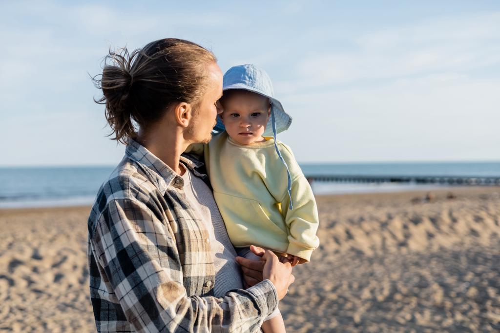 Довговолосий батько дивиться на дівчинку в панамському капелюсі на пляжі в Італії.  - Фото, зображення